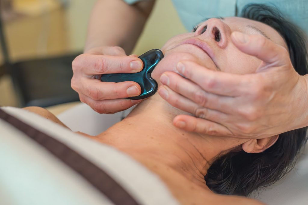 Mittels eines schwarzen Gua Sha Steins massiert Susan Mehr die Kinnpartie einer Kundin während der Gesichts- und Kopfmassage