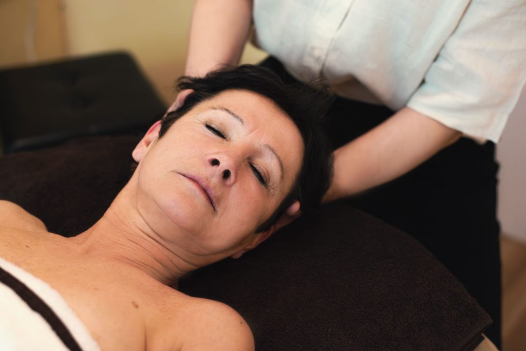Susan Mehr löst Verspannungen im Nacken einer auf dem Rücken liegenden Kundin durch sanftes Ziehen und Drehen des Kopfes während der Gesichts- und Kopfmassage