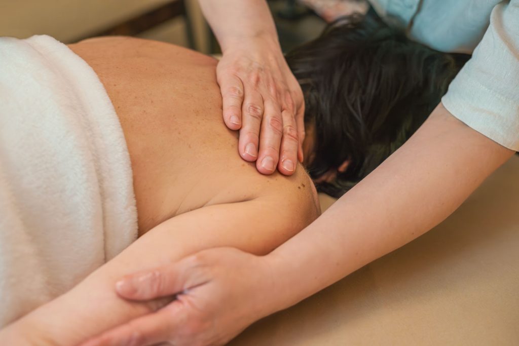 Mit sanften Bewegungen und Druck massiert Susan Mehr die Schulter und den Oberarm einer auf dem Bauch liegenden Kundin während der Teilkörpermassage