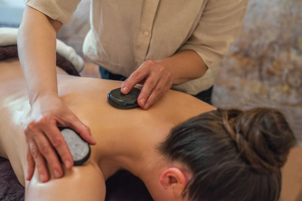 Mit zwei angewärmten Steinen wird der Nacken- und Schulterbereich einer Kundin, die auf dem Rücken liegt, als Teil der Hot-Stone-Massage massiert