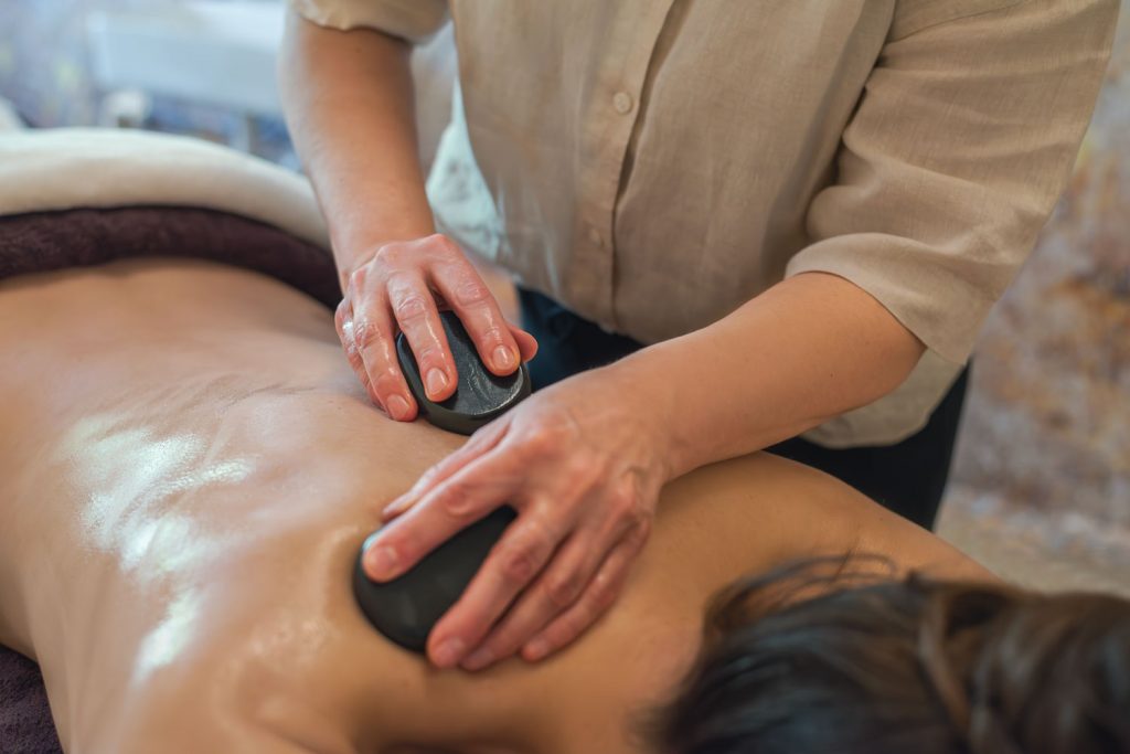 Susan Mehr massiert mit zwei großen warmen Steinen und Massageöl den Rücken einer Kundin in sanften Streichbewegungen im Rahmen der Hot-Stone-Massage