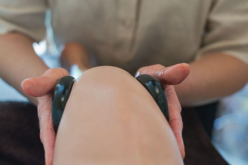 Susan Mehr behandelt das angewinkelte Knie einer Kundin mittels zweier warmer schwarzer Steine von außen als Teilbehandlung der Hot-Stone-Massage