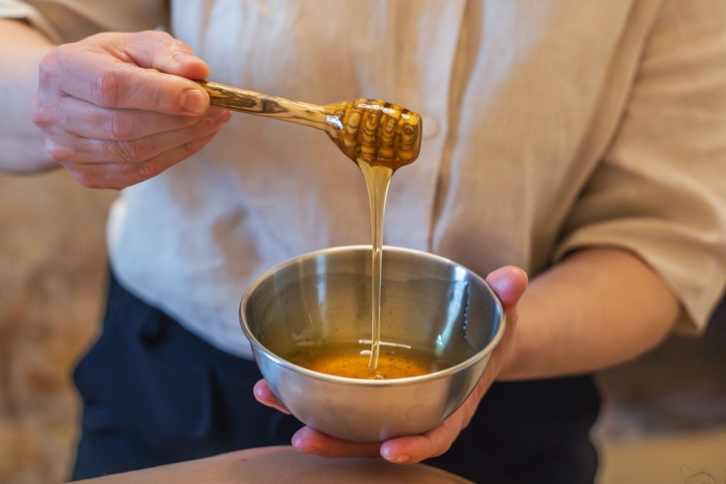 Honig fließt von einem Holzlöffel in eine kleine Metallschale, beides wird von einer Frau in den Händen gehalten