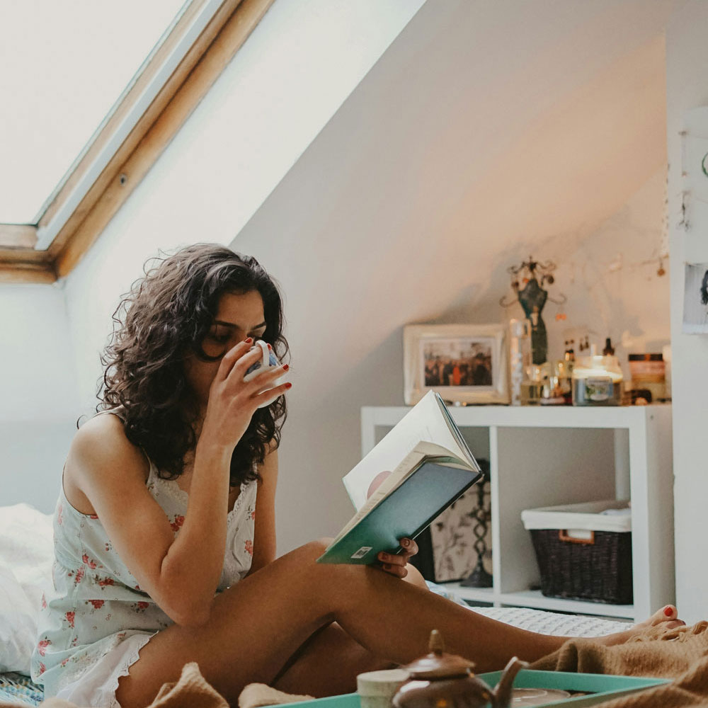 Eine Frau liest sitzend auf ihrem Bett und trinkt dabei Tee aus einer weißen Tasse mit blauen Blumenornamenten